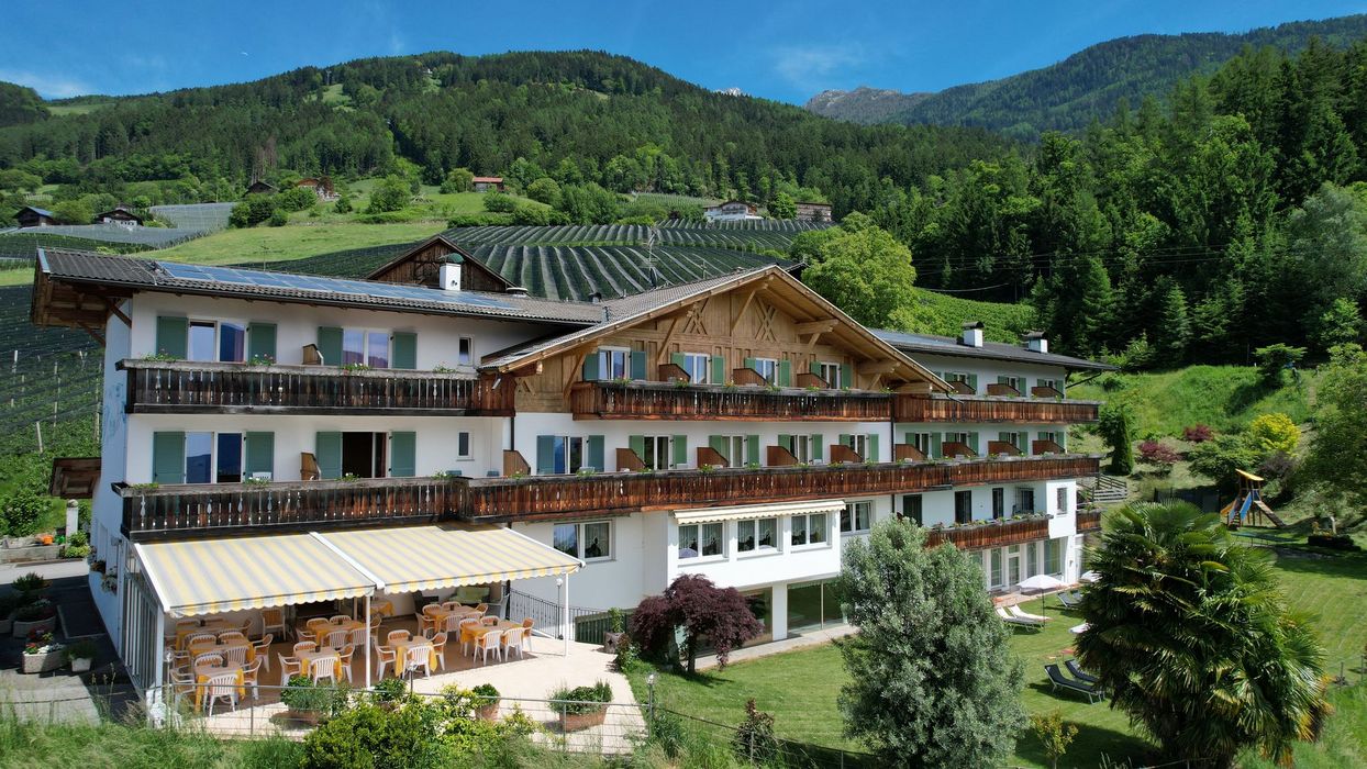 Ein großes, traditionelles Gebäude im alpenländischen Stil mit Holzbalkonen, einer Terrasse mit gelb-weiß gestreifter Markise und einer grünen Bergkulisse im Hintergrund.