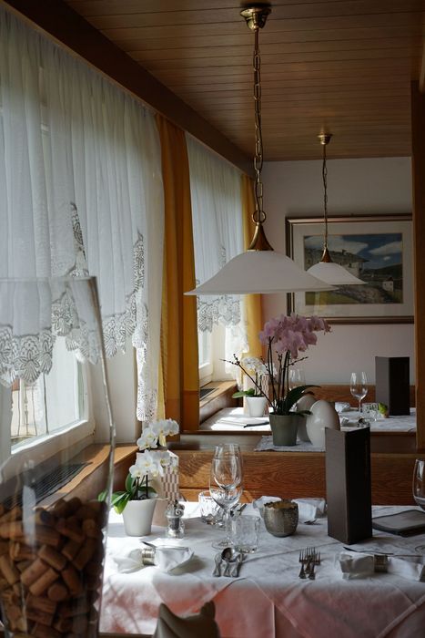 Ein gemütlicher Gastraum mit eingedeckten Tischen, weißen Tischdecken, Orchideen als Tischschmuck und hängenden Lampen vor Fenstern mit Spitzenvorhängen.