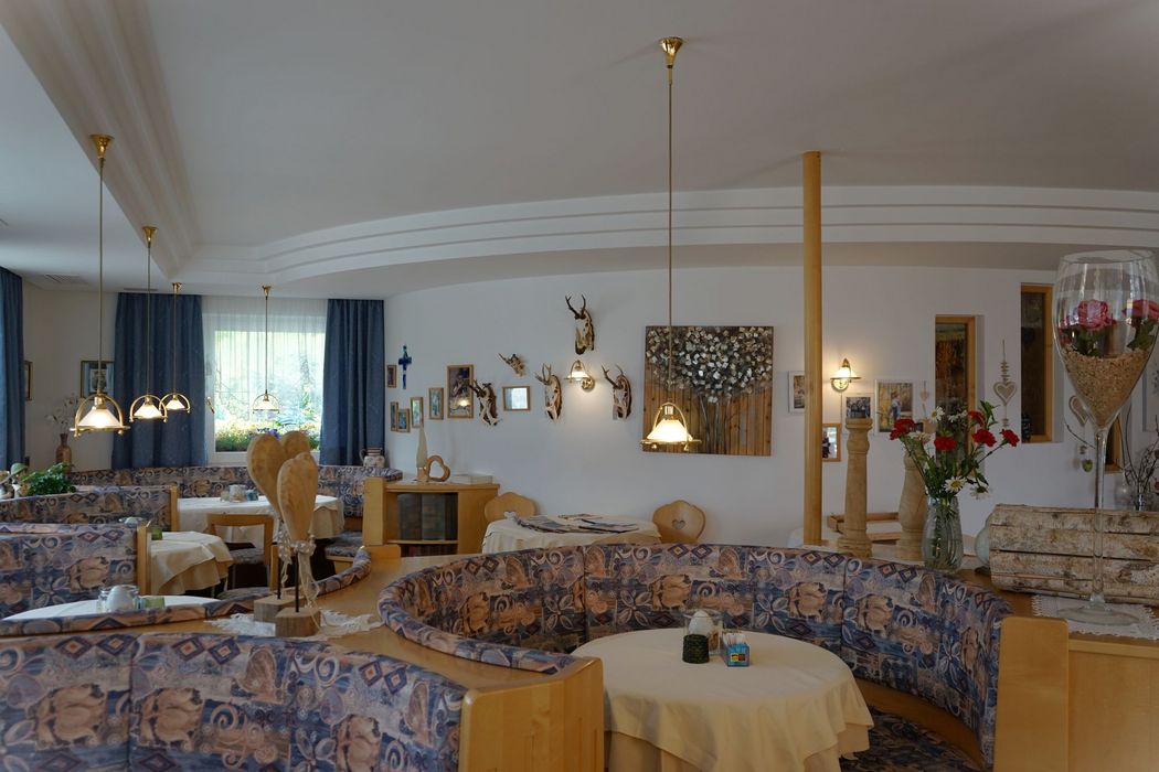 [Translate to italienisch:] Gemütlicher Gastraum mit runder Sitzgruppe, Tisch mit Rosen, großer grüner Vase mit Zweigen und Sonnenblumen, Holzdekoration und Bilder an der Wand