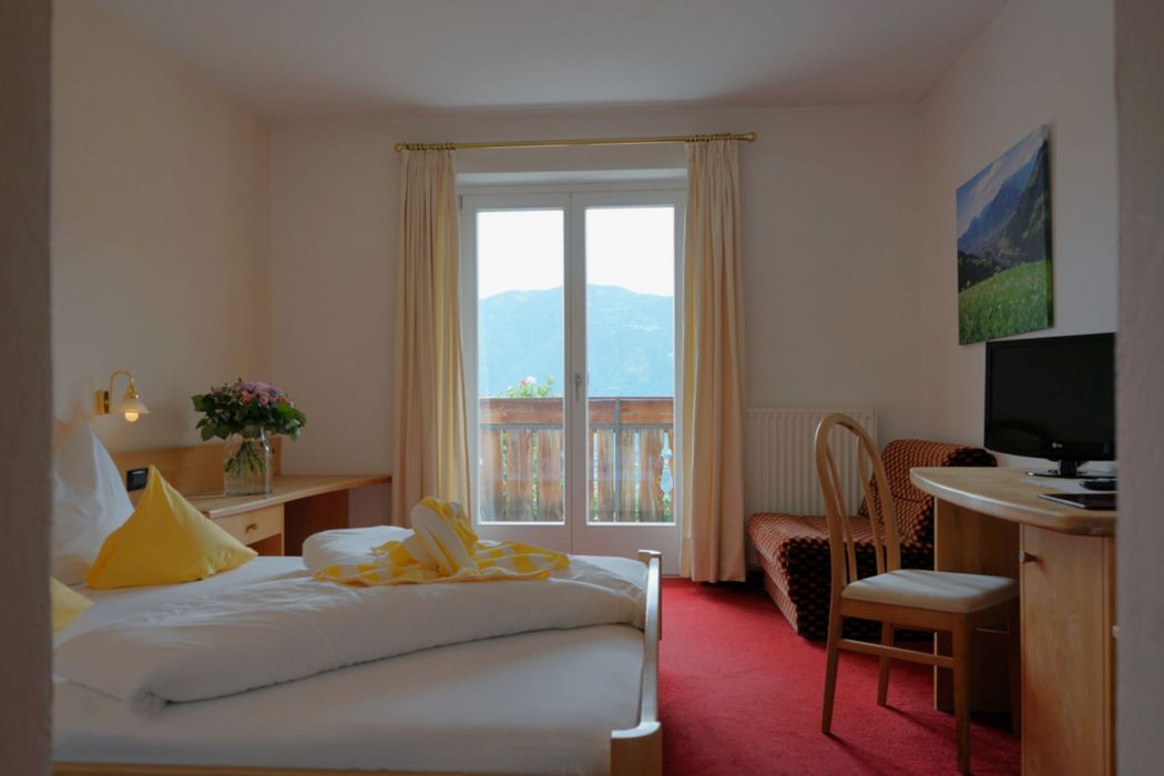 Ein Hotelzimmer mit einem Doppelbett, rotem Teppichboden, einem Schreibtisch mit Stuhl, einem Fernseher und einem Balkonzugang mit Blick auf die Berge.