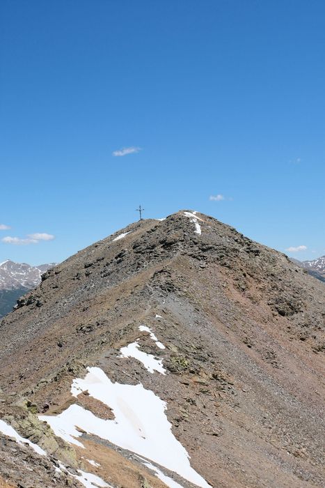 Ein steiler, felsiger Berggipfel mit einem Gipfelkreuz unter strahlend blauem Himmel und vereinzelten Schneeresten am Hang.
