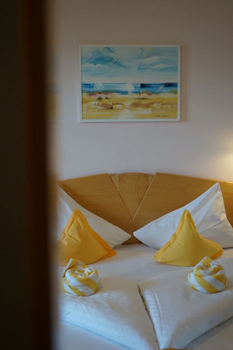 Ein ordentlich gemachtes Hotelbett mit zwei weißen Kopfkissen, zwei gelben Dekokissen und zwei spiralartig gerollten Handtüchern in Weiß und Gelb darauf, an einer Wand hängt ein gerahmtes Gemälde einer Meereslandschaft.