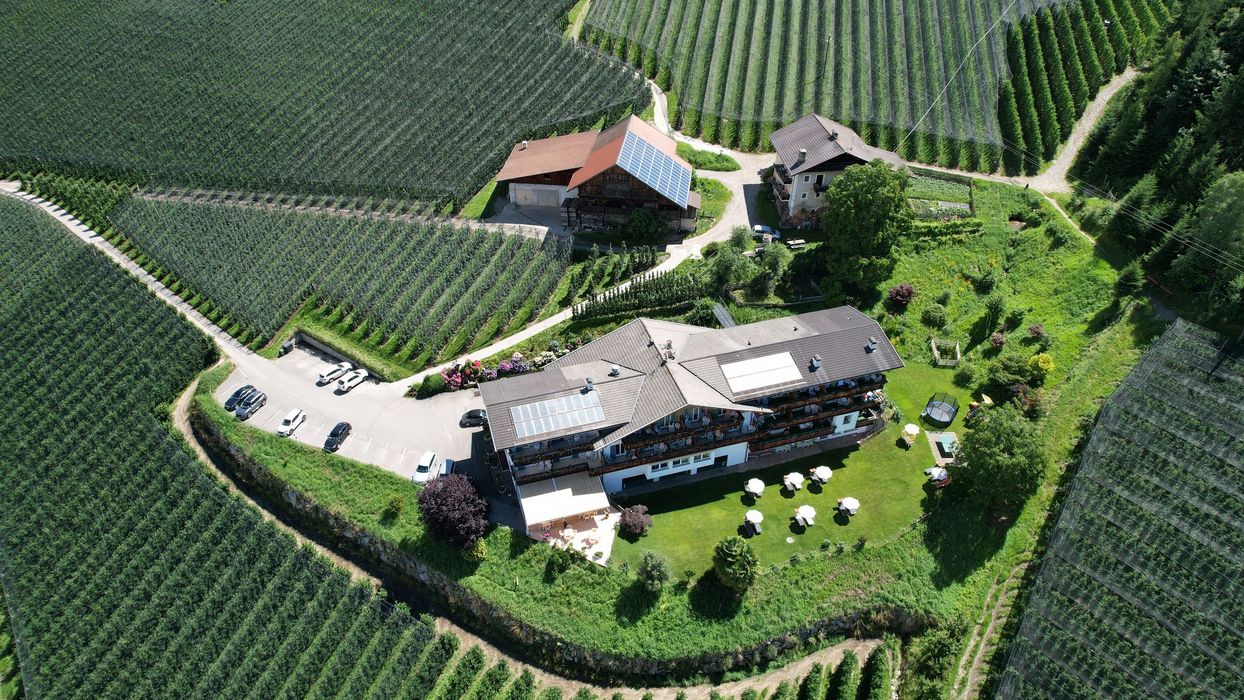 Luftaufnahme eines Hotels mit Parkplatz und Gastgarten, umgeben von weitläufigen Apfelplantagen in hügeliger Landschaft.