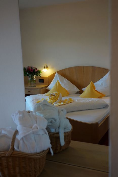 Blick in ein Hotelzimmer mit einem Doppelbett aus hellem Holz, dekoriert mit gelben Kissen und einem gerollten Handtuch, sowie Körbe mit weißen Bademänteln im Vordergrund.