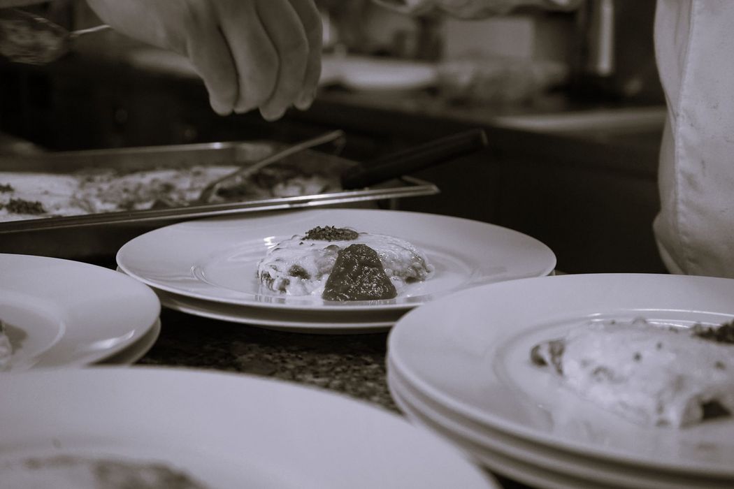 Ein Koch richtet in einer Gastronomieküche auf mehrere weiße Teller kleine Portionen mit Käse überbackener Speisen und Tomatensoße an.