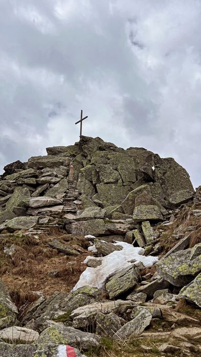 Ein steiler, felsiger Pfad führt auf einen Berggipfel, der mit einem Gipfelkreuz und vereinzelten Schneeresten bedeckt ist, vor einem bewölkten Himmel.