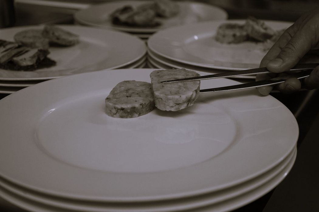 Eine Hand platziert mit einer Pinzette eine Scheibe Wurst oder Fleisch auf einen weißen Teller, im Hintergrund stehen weitere Teller für die Anrichtung bereit.