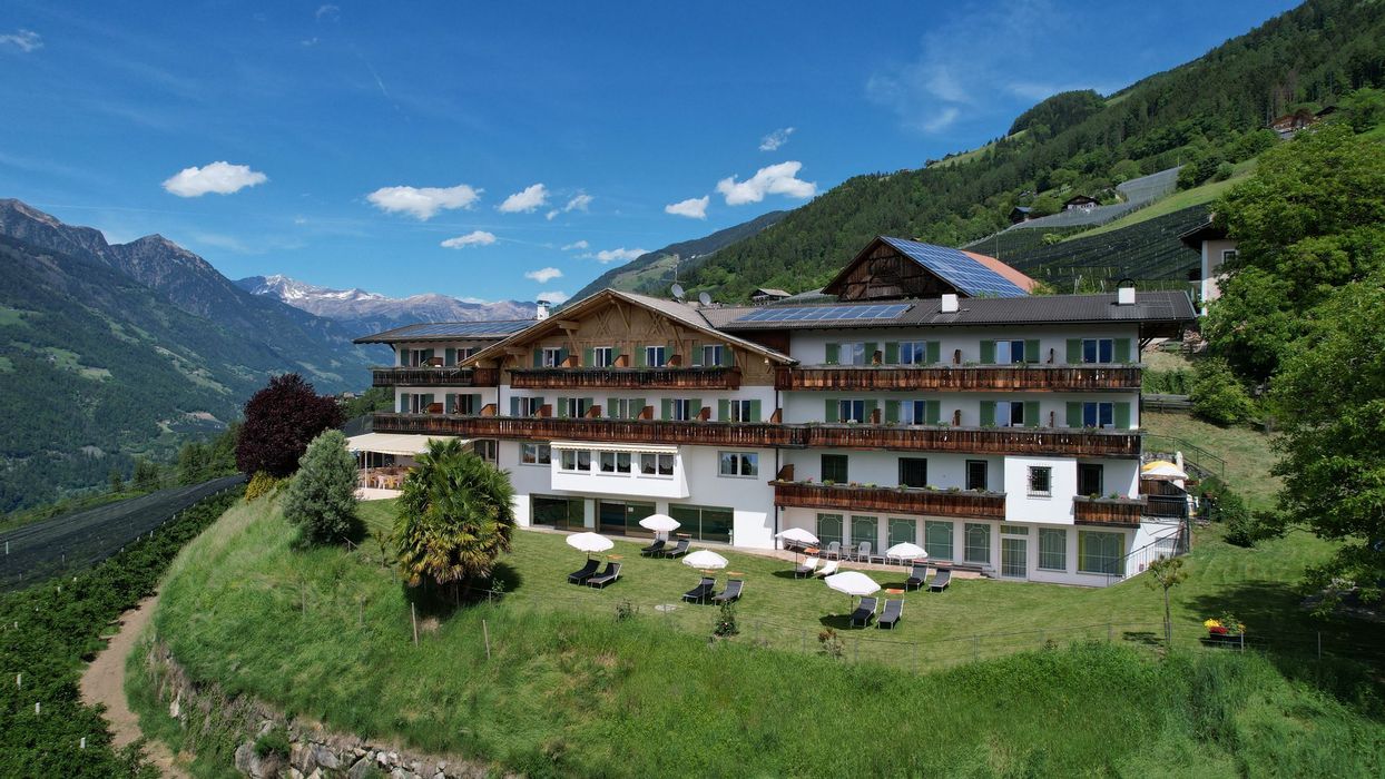 Ein großes, traditionelles alpenländisches Hotelgebäude mit Holzbalkonen vor einer Berglandschaft mit einer Liegewiese und Sonnenschirmen im Vordergrund.