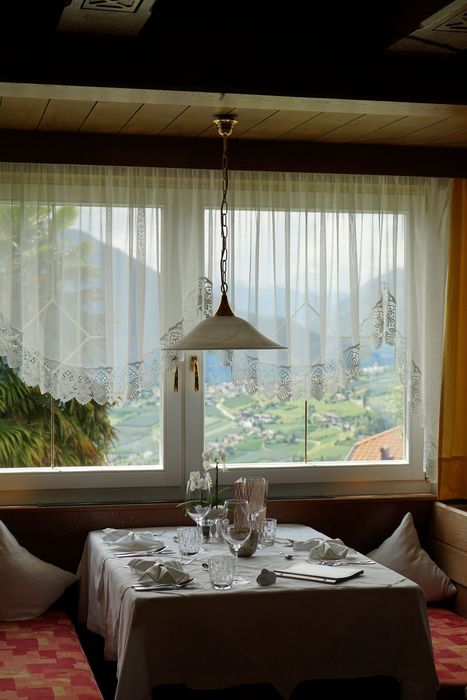 Ein gedeckter Tisch für zwei Personen vor einem Fenster mit weißem Spitzengardinen-Vorhang, durch das man auf ein Tal in den Bergen blickt.