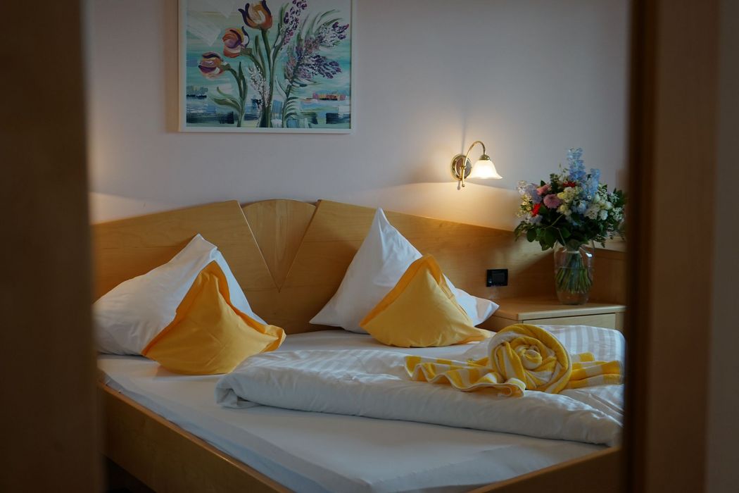 Ein gemütliches Hotelbett mit einem hölzernen Kopfteil, weißen Bettbezügen, gelben Dekokissen, einem strauß Blumen und einem handtuch in Form einer Blüte.