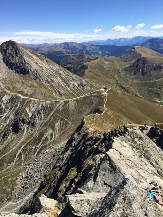 Ein Blick von einem felsigen Berggipfel über eine alpine Landschaft mit geschwungenen Wanderwegen, schroffen Hängen und einer kleinen Schutzhütte in der Ferne bei strahlend blauem Himmel.