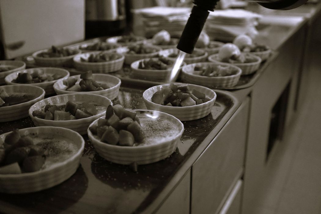 Eine Nahaufnahme in Schwarz-Weiß zeigt, wie eine Hand mit einem Flambierbrenner eine Reihe kleiner Förmchen bearbeitet, die mit Erdbeerstückchen gefüllt sind.