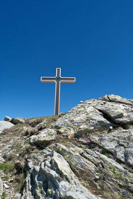 Ein Gipfelkreuz aus Holz auf einem felsigen Berggipfel vor einem wolkenlosen, tiefblauen Himmel.