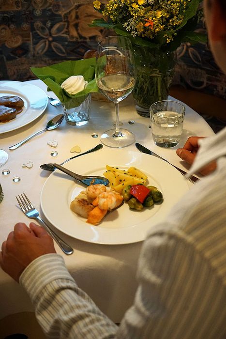 Eine Person sitzt an einem fein gedeckten Tisch mit einem Teller, auf dem gebratener Fisch, Garnelen, Kartoffeln und Rosenkohl serviert werden, ergänzt durch ein Weinglas und eine Blumenvase.