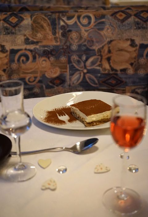 Ein Stück Tiramisu auf einem weißen Teller mit einem Gabelabdruck im Kakaopulver, dazu zwei Weingläser und kleine herzförmige Dekorationen auf einem gedeckten Tisch.