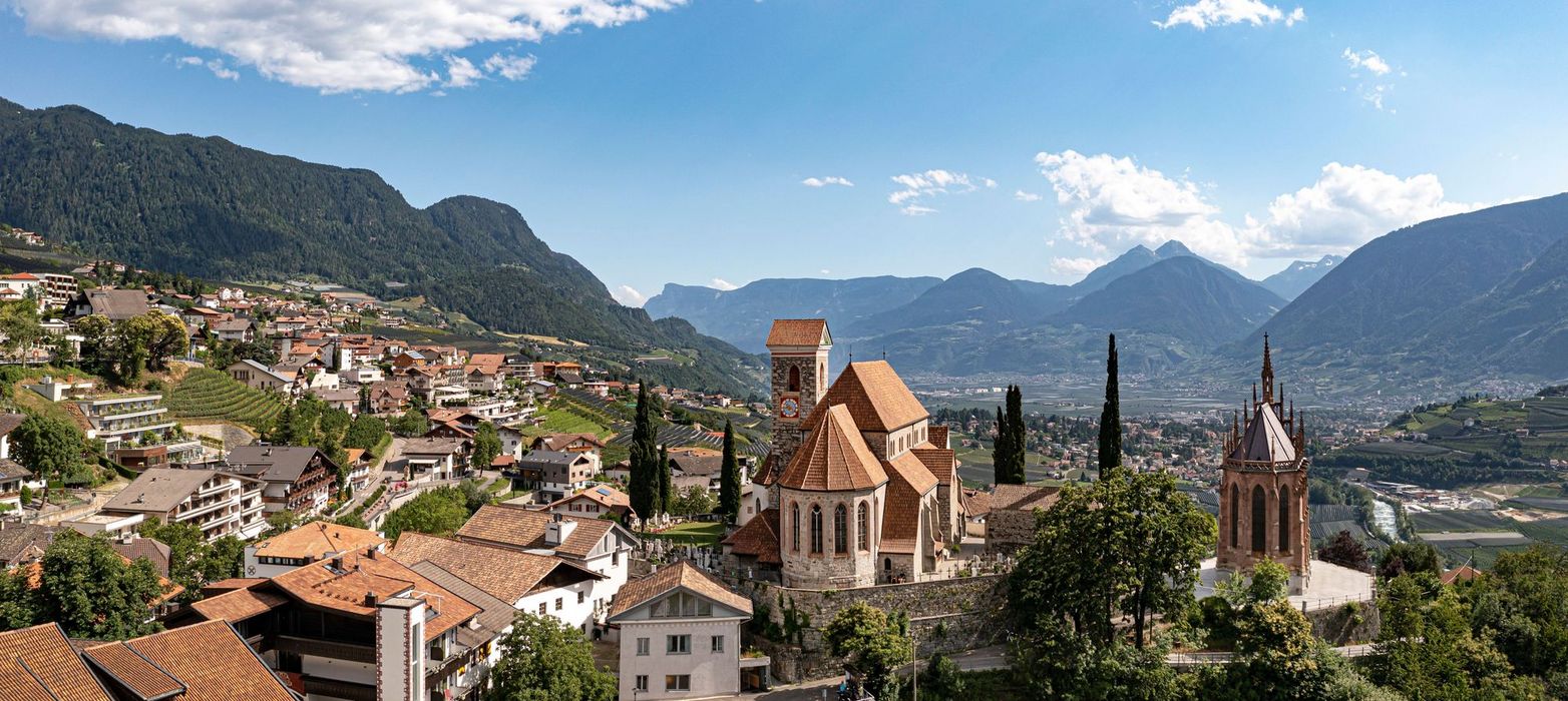 Ein Panoramablick auf die Kirche St. Nikolaus in Dorf Tirol vor einer Kulisse aus Bergen mit Weinbergen und einem Tal im Hintergrund.