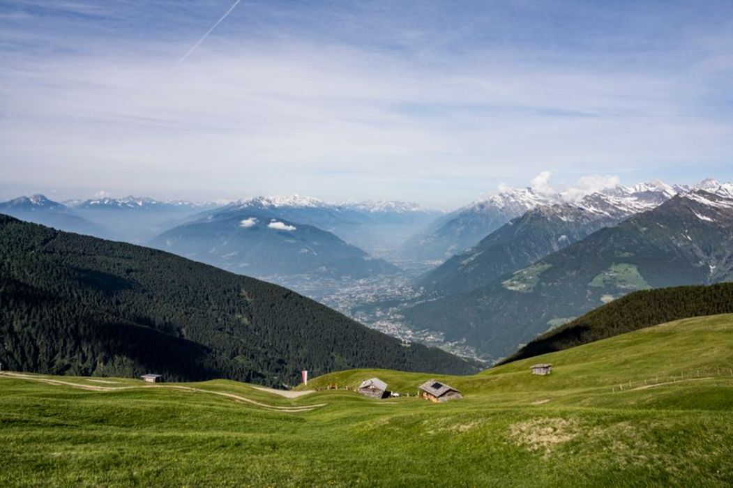 Weite Berglandschaft mit grünen Almwiesen und kleinen Holzstadln im Vordergrund sowie einem Blick in ein tiefes Tal mit schneebedeckten Gipfeln am Horizont.