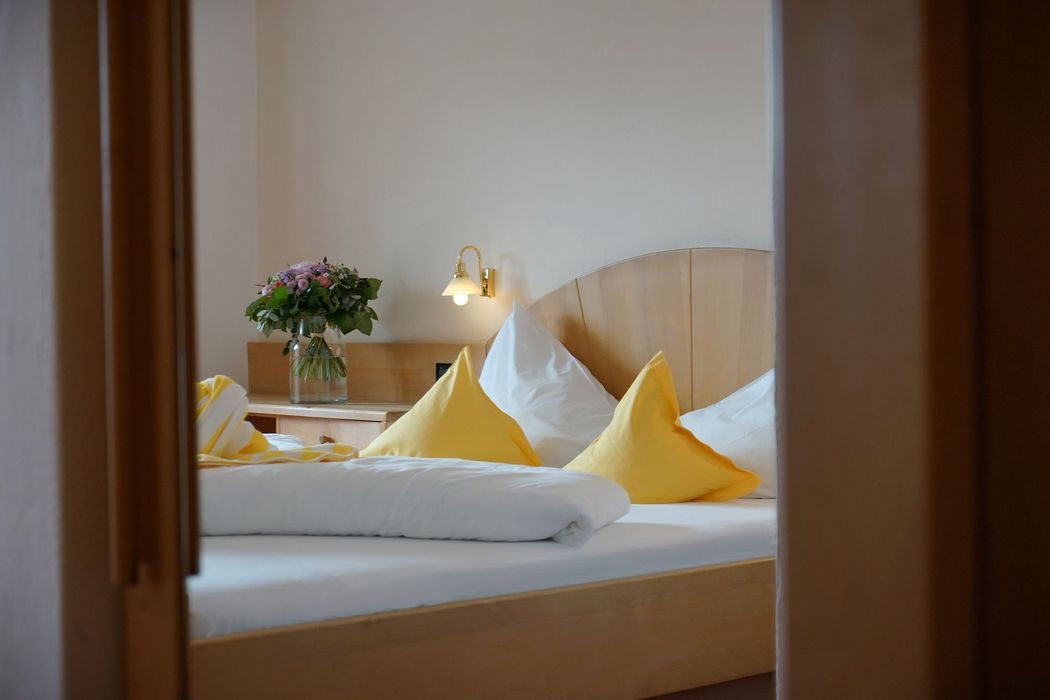 Blick durch einen Türrahmen in ein Schlafzimmer mit einem Doppelbett aus hellem Holz, bezogen mit weißer Bettwäsche und leuchtend gelben Zierkissen sowie einem Blumenstrauß auf dem Nachttisch.