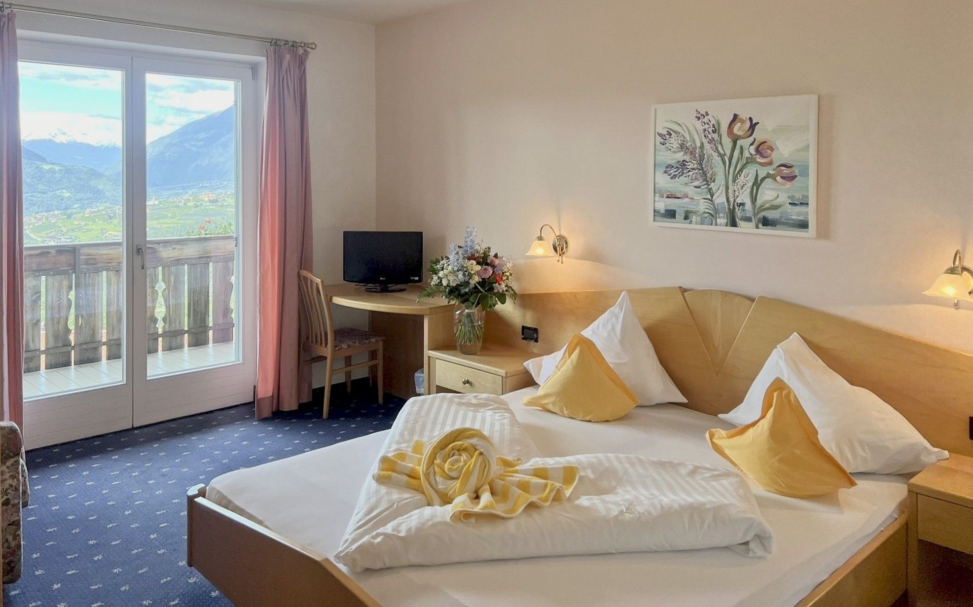 Helles Hotelzimmer mit Doppelbett, gelben Kissen, Blumenstrauß, kleinem TV und Balkon mit Bergblick