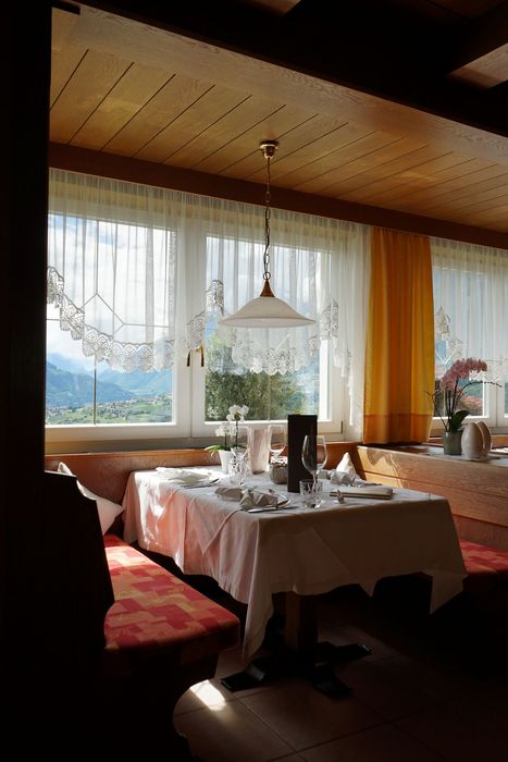 Ein gemütlicher, mit einer weißen Tischdecke gedeckter Tisch in einer traditionellen Holzstube steht vor einem Fenster mit Blick auf eine sonnige Berglandschaft