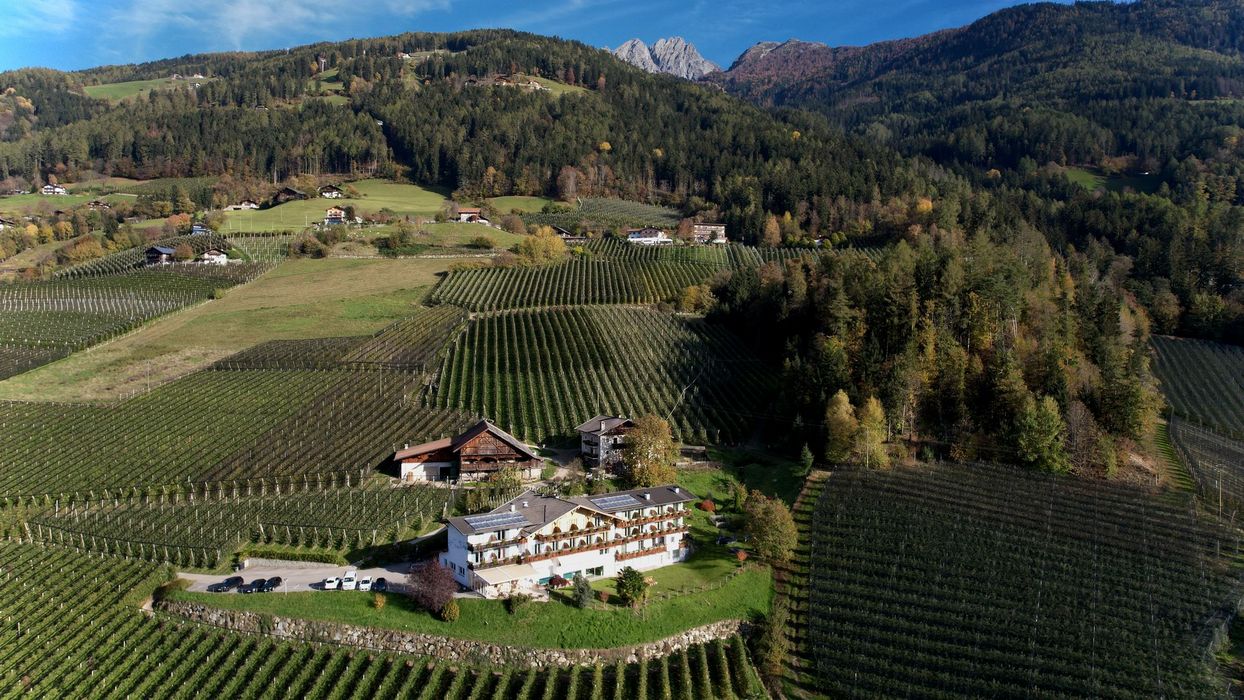 Ein von oben aufgenommenes Foto eines Hotels inmitten weitläufiger Apfelplantagen an einem sonnigen Hang mit bewaldeten Bergen und Felsen im Hintergrund.