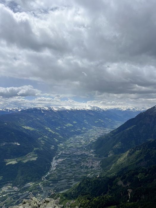 Blick von einem Berg auf ein tief gelegenes Tal mit Feldern und kleinen Ortschaften, umrahmt von hohen Bergen mit schneebedeckten Gipfeln unter einem bewölkten Himmel.