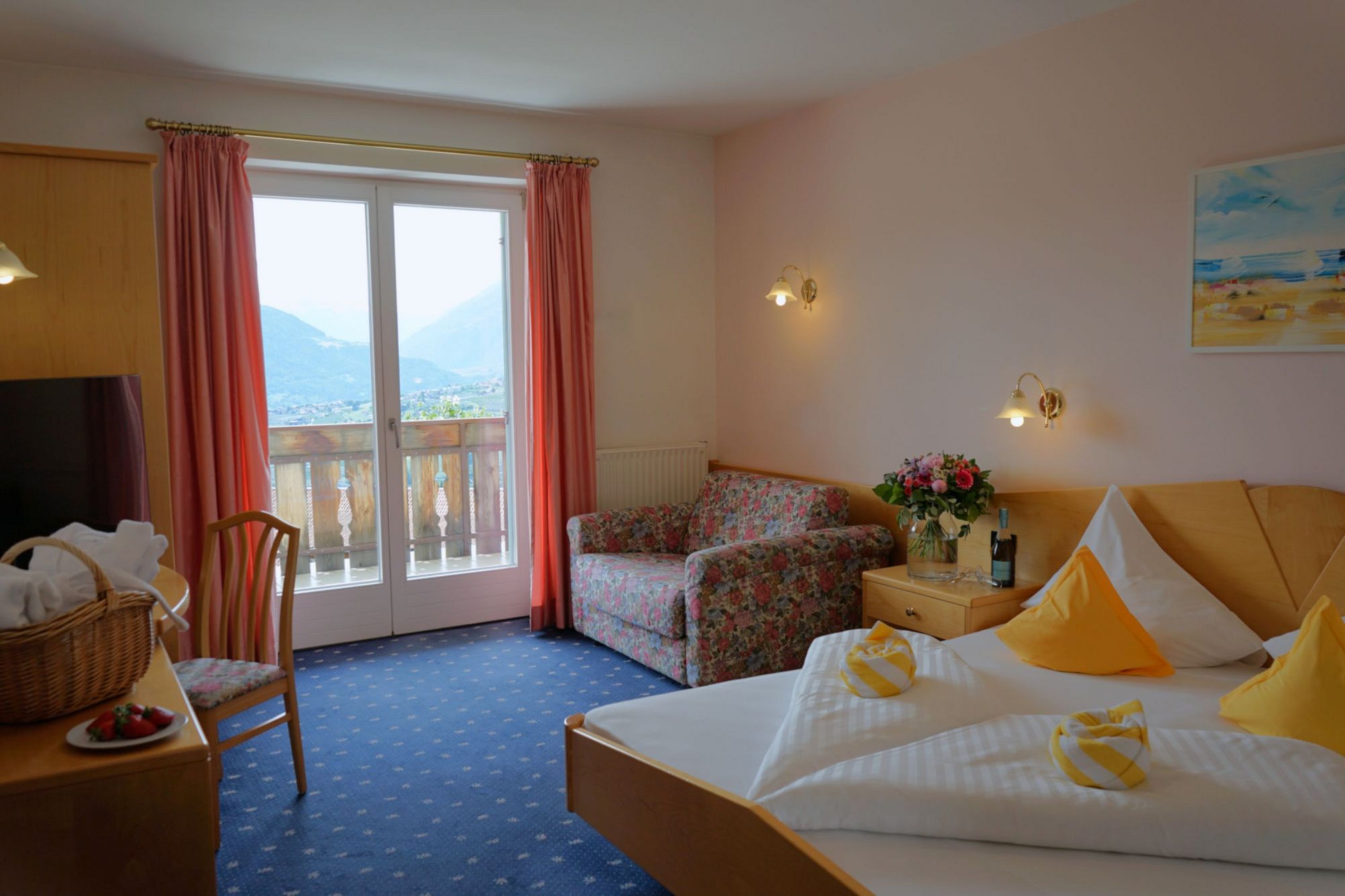 [Translate to italienisch:] Gemütliches Hotelzimmer mit Doppelbett, Blumenstrauß, TV, bequemer Sitzgelegenheit und Balkon mit Bergblick