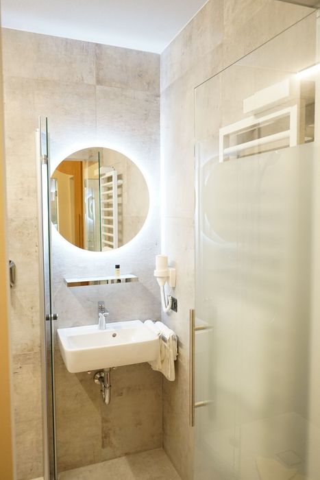 Ein modernes Badezimmer mit grauen Fliesen, einem weißen Waschbecken, einem beleuchteten runden Spiegel und einer angrenzenden Dusche mit Glastür.