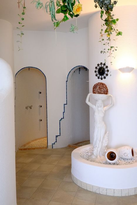 Ein Wellnessbereich mit zwei gefliesten Duschkabinen, die durch Torbögen zugänglich sind, sowie einer weißen Frauenstatue in einem mit Kieselsteinen gefüllten Brunnenbecken, dekoriert mit Rankpflanzen von der Decke.